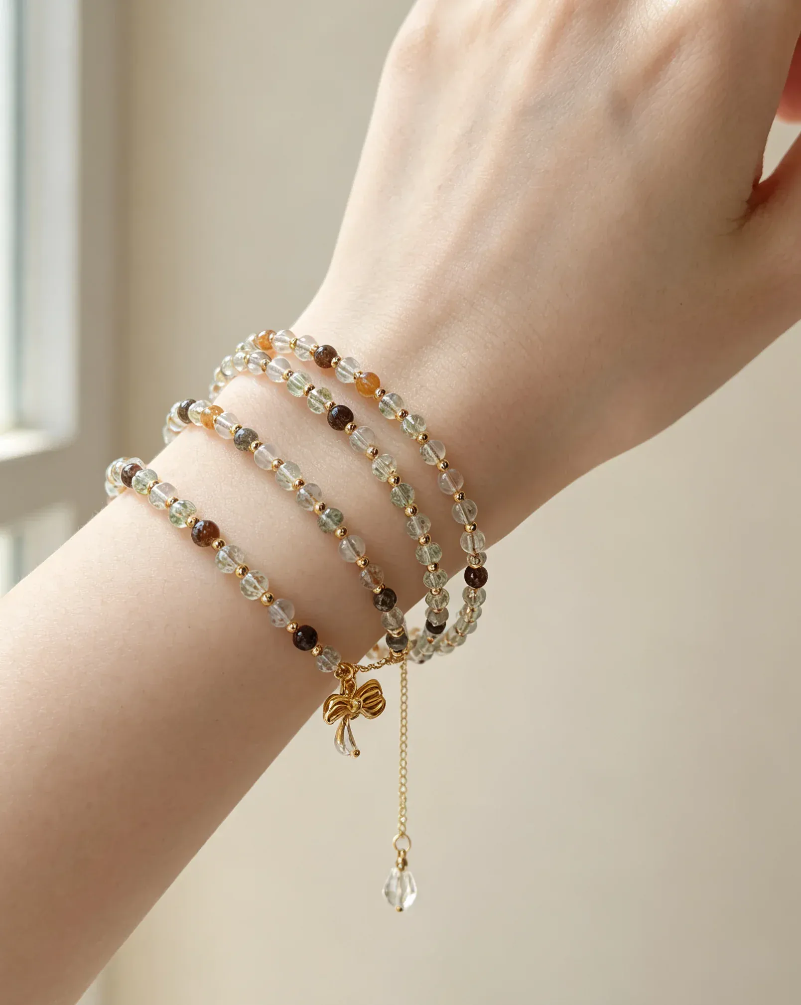 Hand wearing layered natural crystal bracelets with gold bow charm, genuine gemstone healing accessories for women